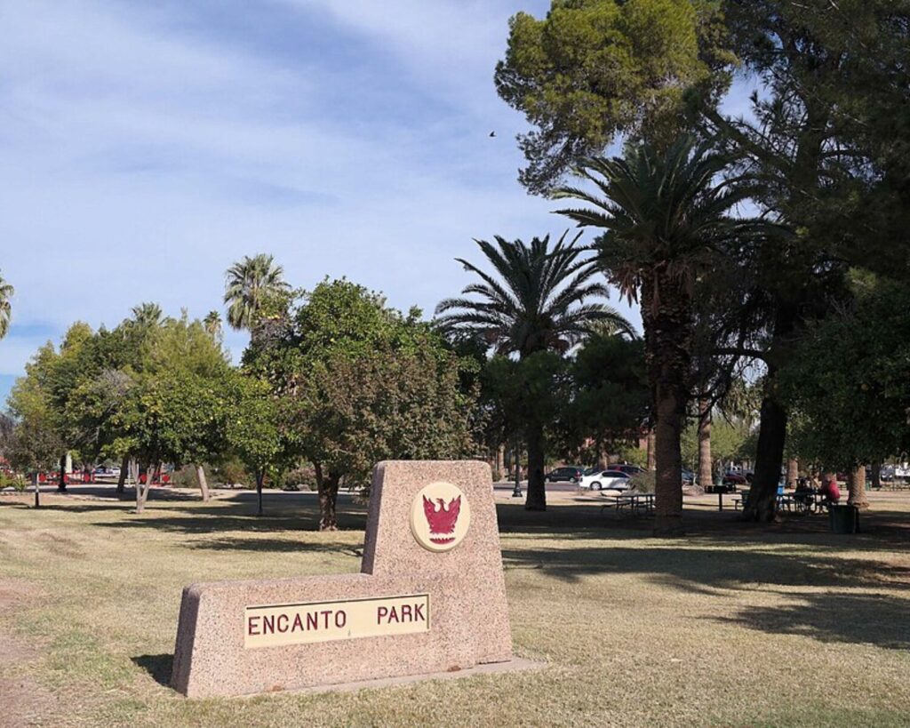 Encanto Park entrance sign on a grassy lawn surrounded by palm trees and greenery under a clear sky.