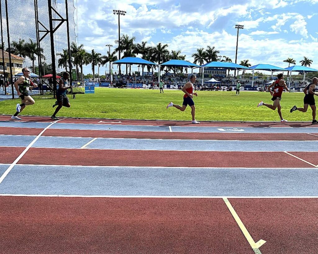 Athletes running on a track during the 2022 National Senior Games 800 meter race with spectators and palm trees in the background.