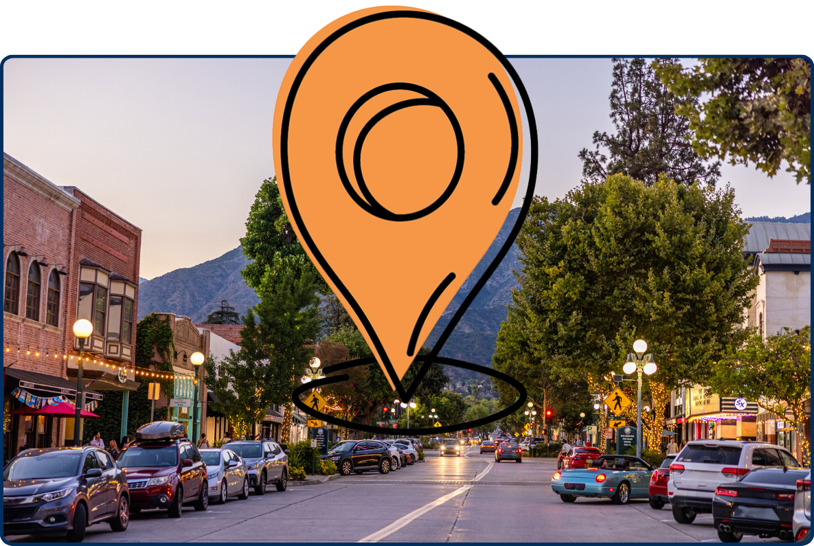 Downtown Monrovia, California, at dusk, with parked cars, lit storefronts, tree-lined streets, and the San Gabriel Mountains in the background.