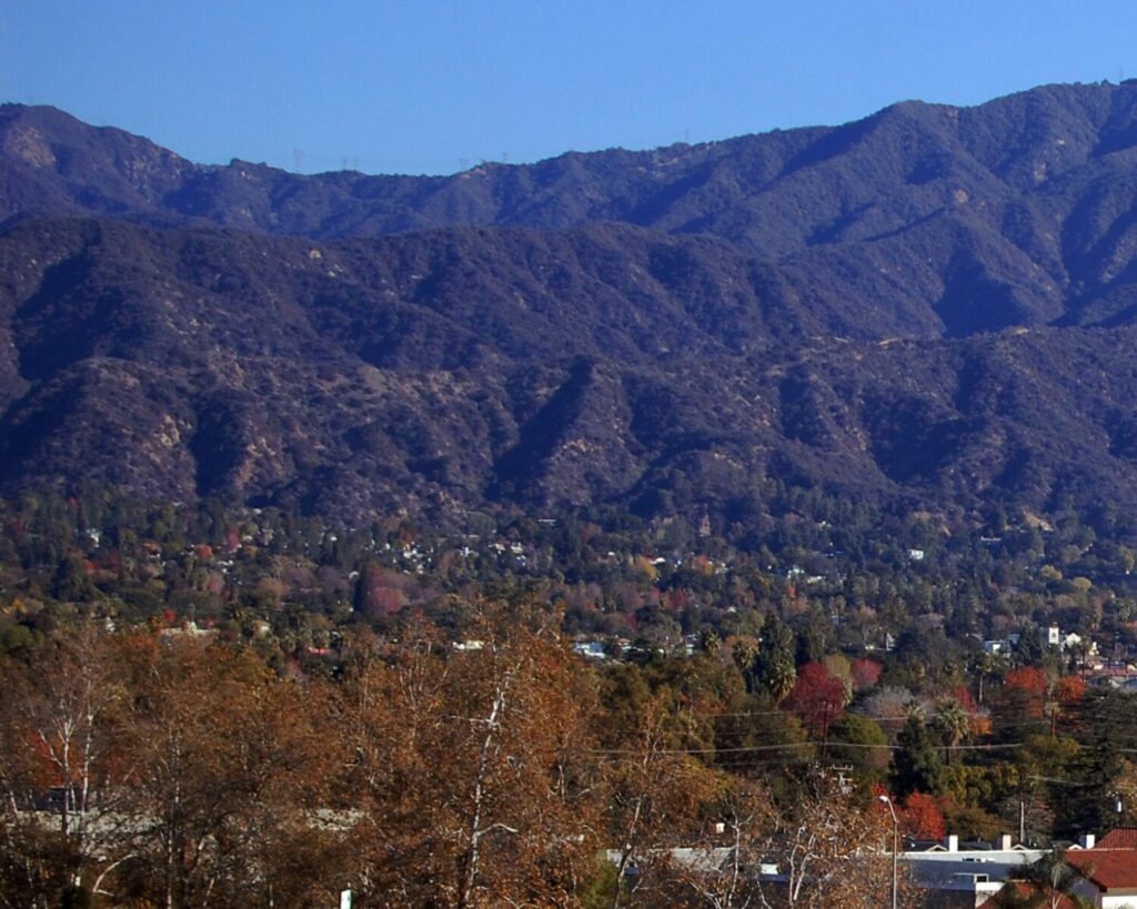 Mountain range rising behind a town, with autumn-colored trees in the foreground and a clear blue sky above.