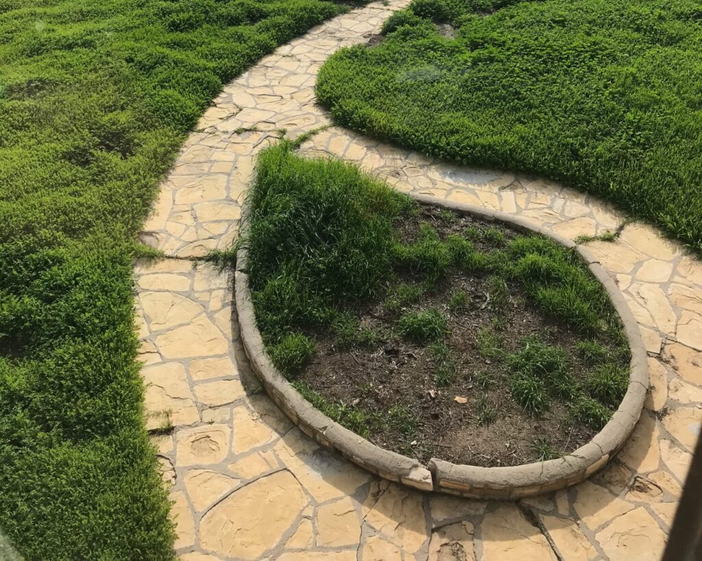 Curved stone pathway leading through green grass to an outdoor pool surrounded by trees and distant hills.