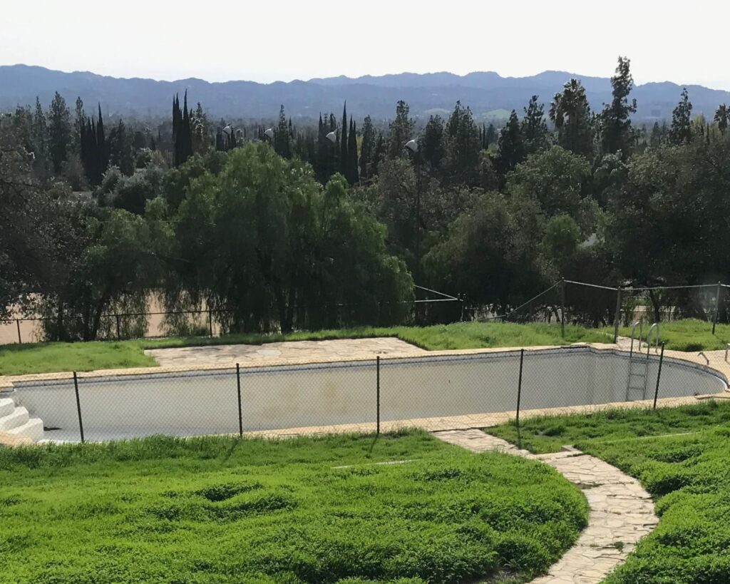 Curved stone pathway leading through green grass to an outdoor pool surrounded by trees and distant hills.