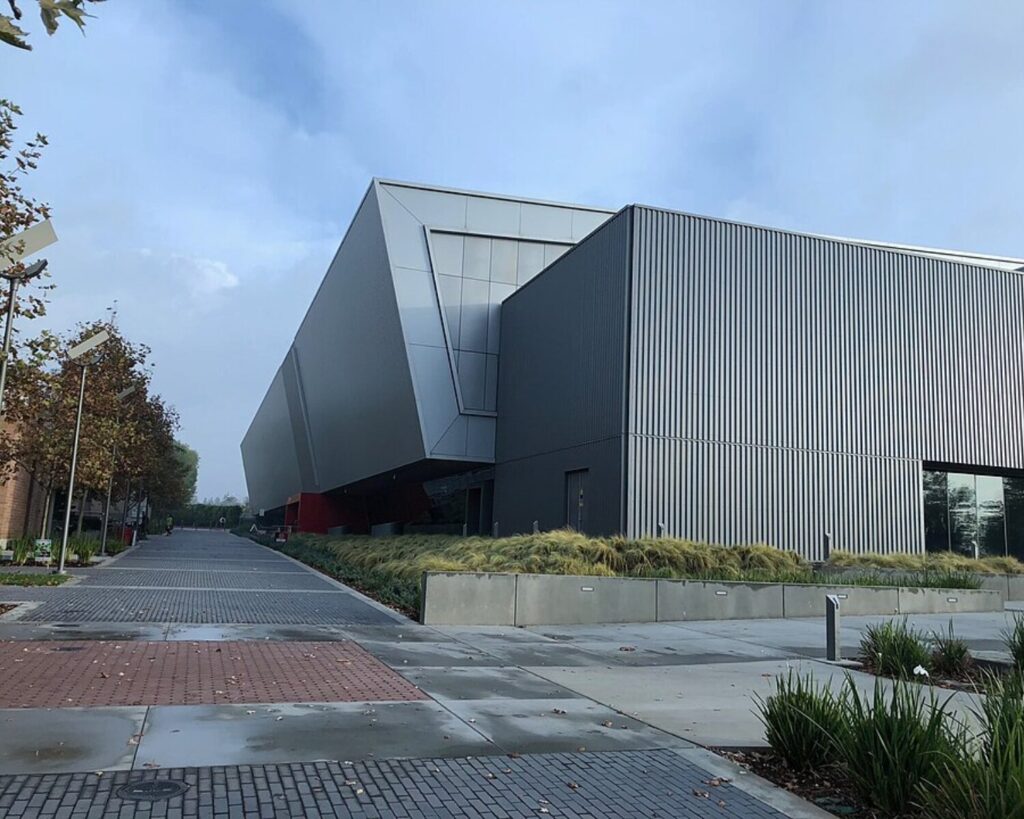 Modern angular building with gray metal panels and a slanted facade beside a paved walkway.