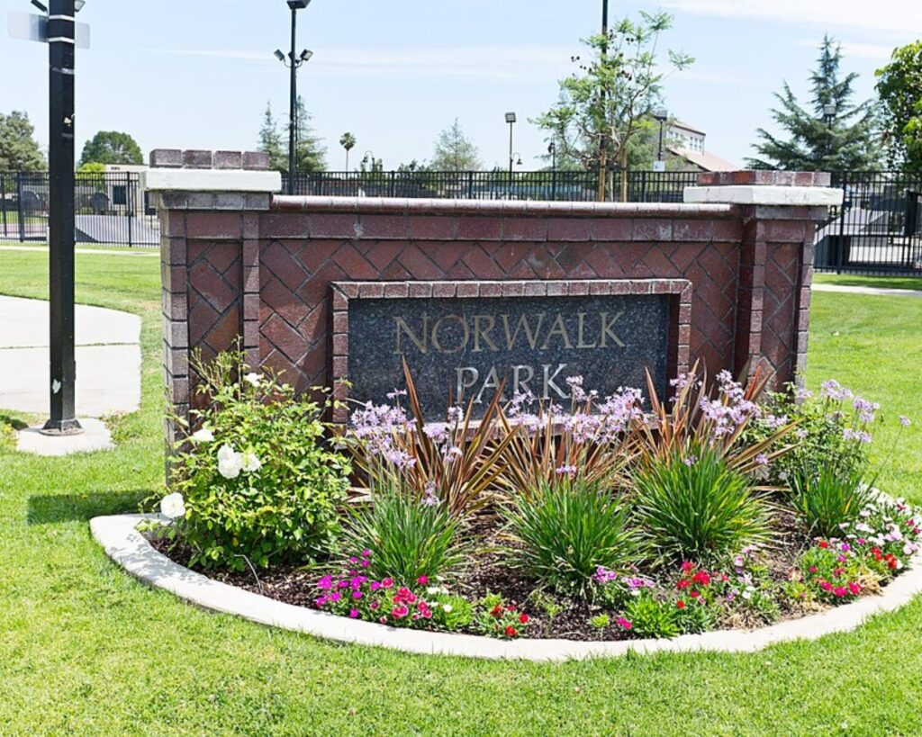 Brick sign reading 'NORWALK PARK' set in a brick planter, with a colorful flower bed in front and a green lawn surrounding it.