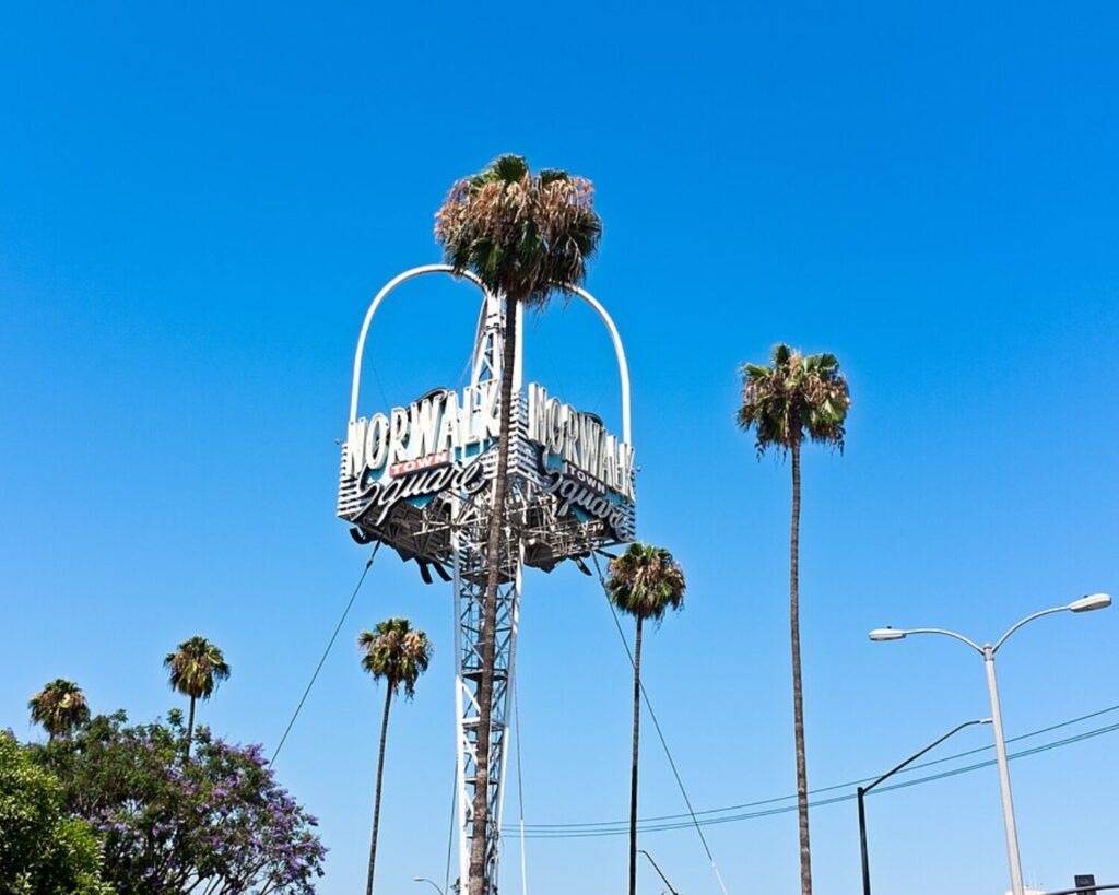 Norwalk Town Square sign on a tall metal structure with palm trees against a bright blue sky, sunny day.