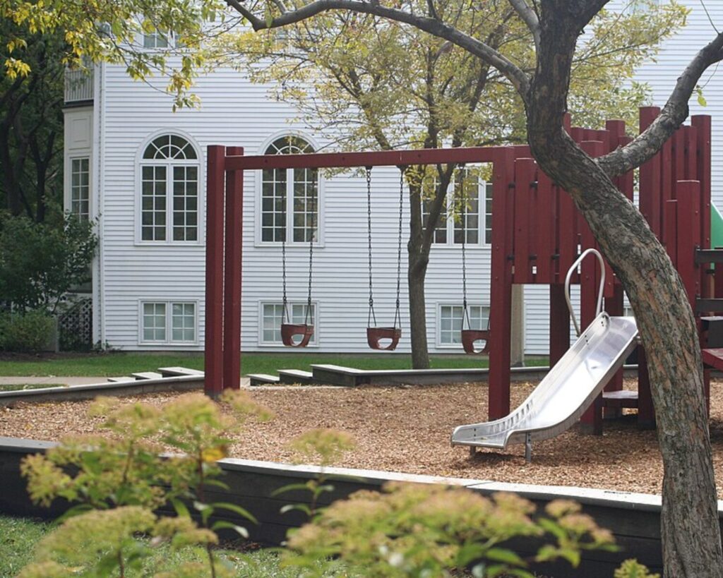 Children’s playground in Bauler Park featuring swings and a slide, surrounded by trees and residential buildings in the background.