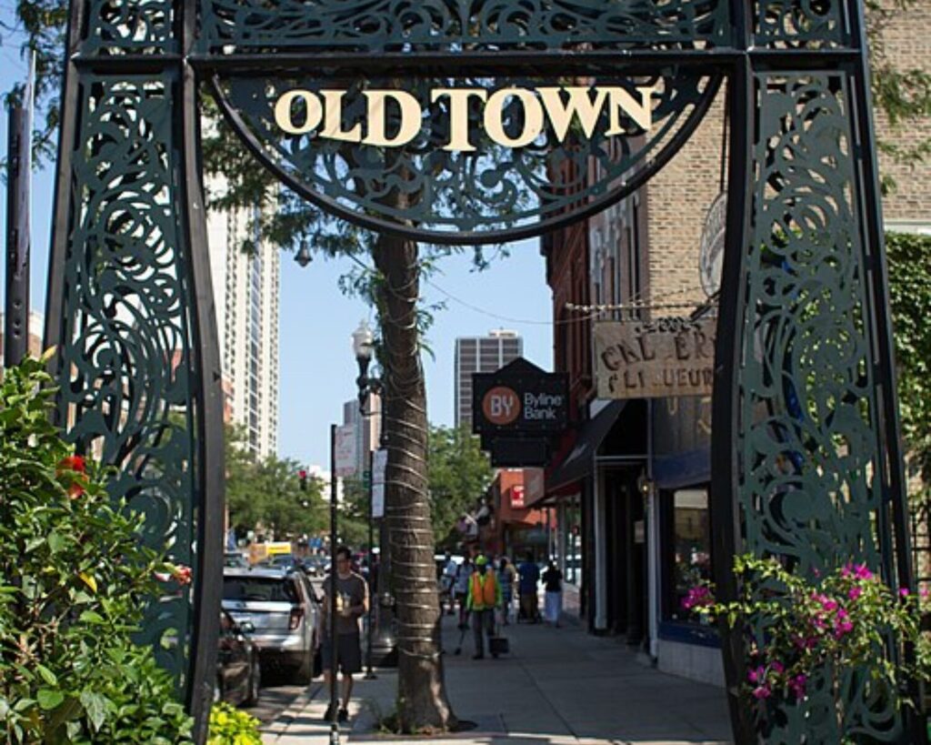 Decorative “Old Town” metal archway sign over a sidewalk in Chicago’s Old Town neighborhood, with shops, trees, and pedestrians along the street.