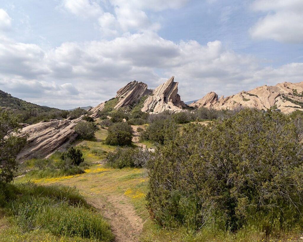 Vasquez Rocks landscape with jagged rock formations, grassy trails, and scattered shrubs under a partly cloudy sky.