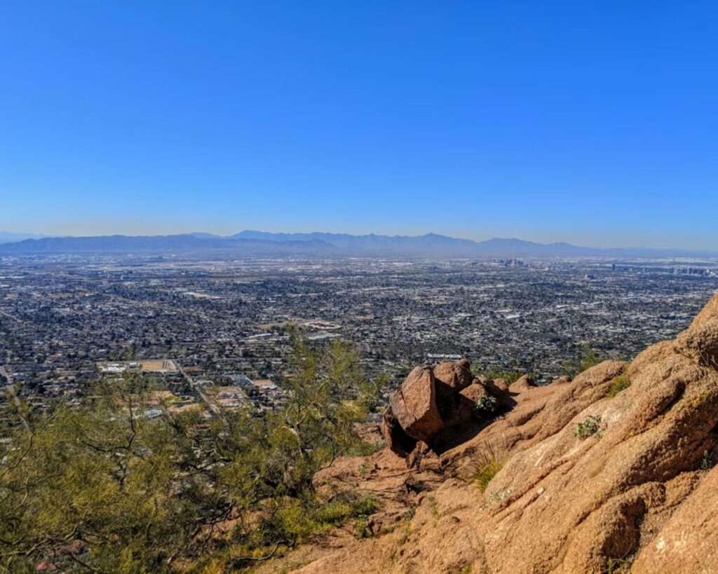 Panoramic aerial view of a sprawling city landscape surrounded by mountains, seen from a rocky hillside under a clear blue sky.