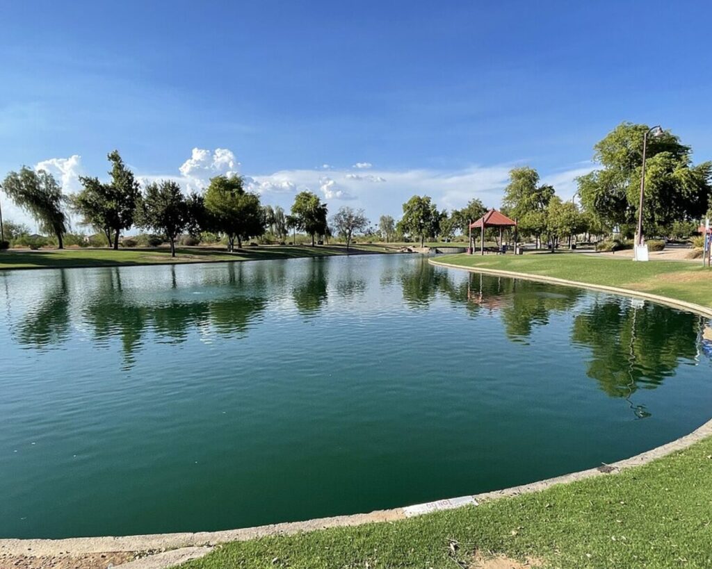 Rio Vista Community Pond with calm water reflecting trees, grassy park area, and a small pavilion under a clear blue sky.