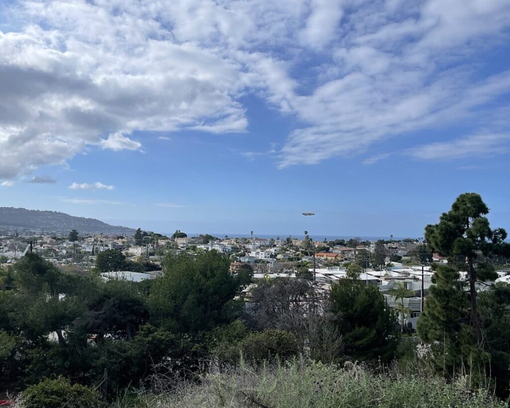 Goodyear blimp flying low over Torrance with residential area, trees, and ocean in the background under a partly cloudy sky.