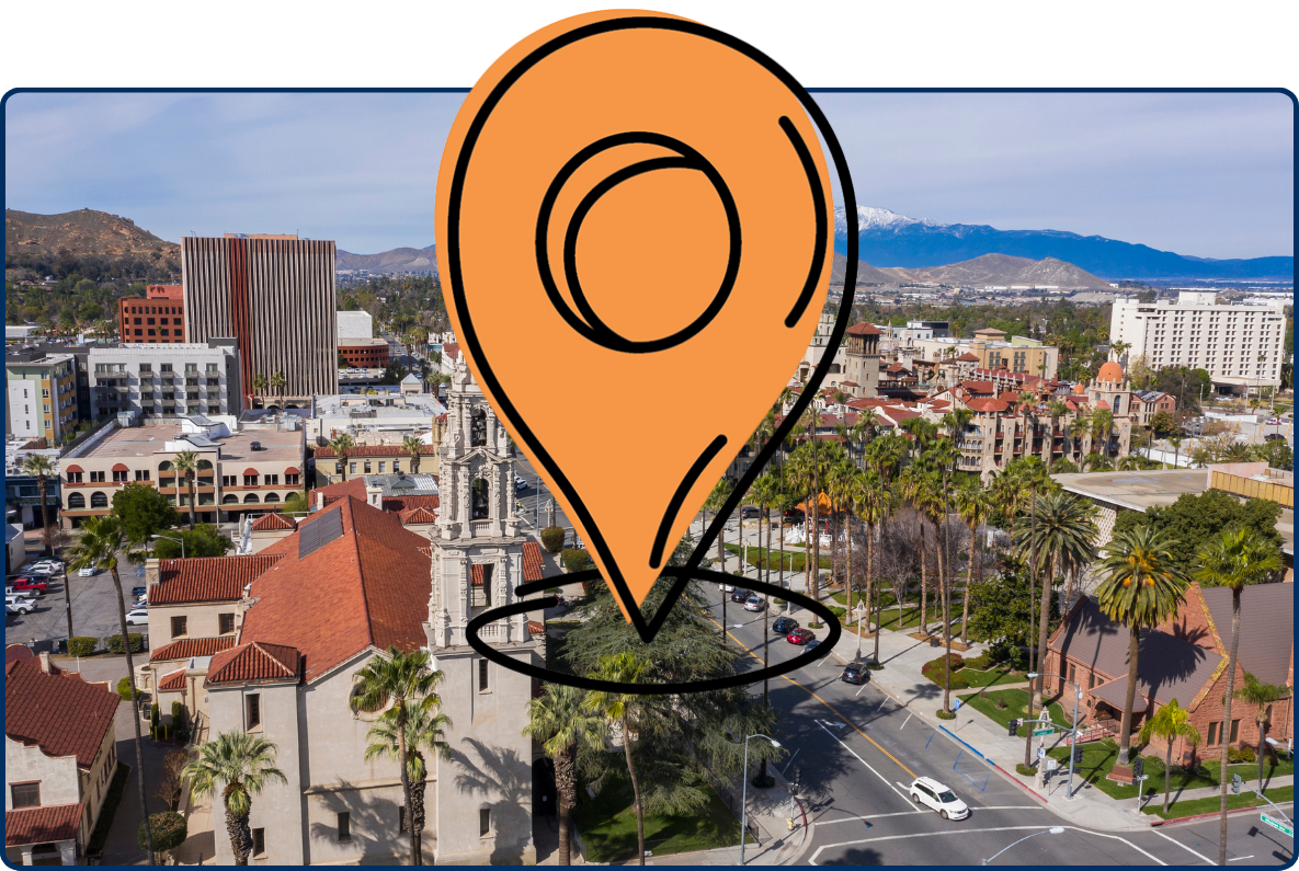 Aerial view of downtown Riverside, California, featuring palm-lined streets, historic buildings with red-tiled roofs, modern hotels, and snow-capped mountains in the background.