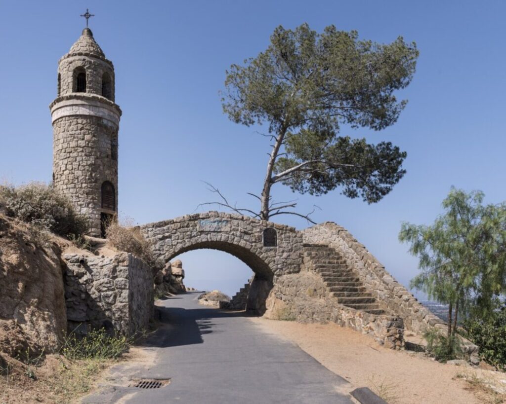 Stone tower and arched bridge on Mount Rubidoux in Riverside, California, overlooking a paved hillside path with sparse trees under a clear blue sky.
