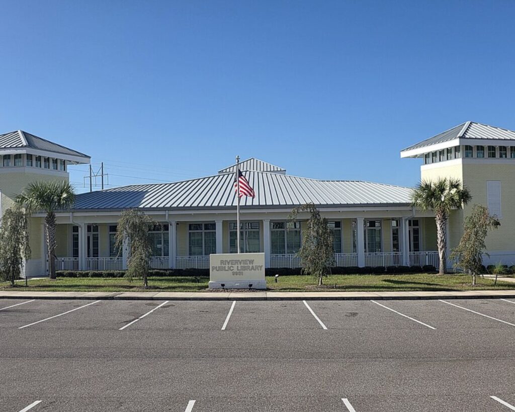 Front view of Riverview Public Library: a pale yellow, one-story building with a metal roof, white columns, and an American flag on a central flagpole; palm trees and a manicured lawn in front.