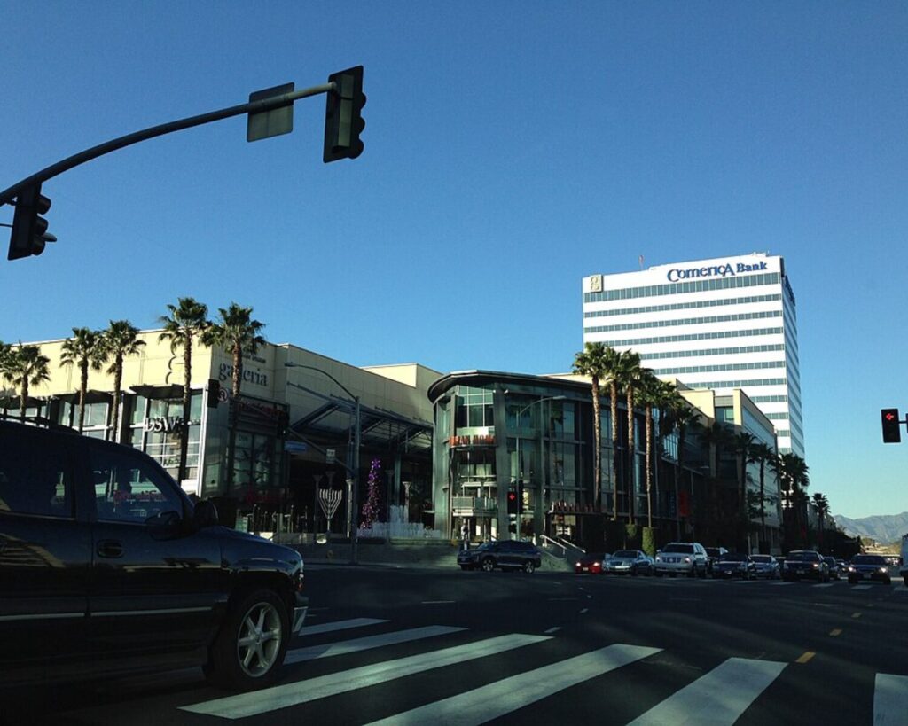 Busy intersection in Sherman Oaks, Los Angeles, with palm-lined shopping buildings, traffic lights, cars, and office towers under a clear sky.