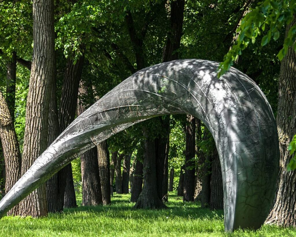 Large curved metallic sculpture bending through a green, sunlit forest in a park setting