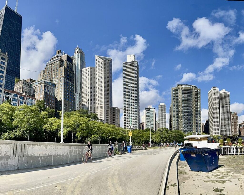 Scenic view of the Chicago Lakefront Trail along Lake Michigan, showing a paved path for cyclists and pedestrians with green park areas, city skyline in the distance, and a bright partly sunny sky.
