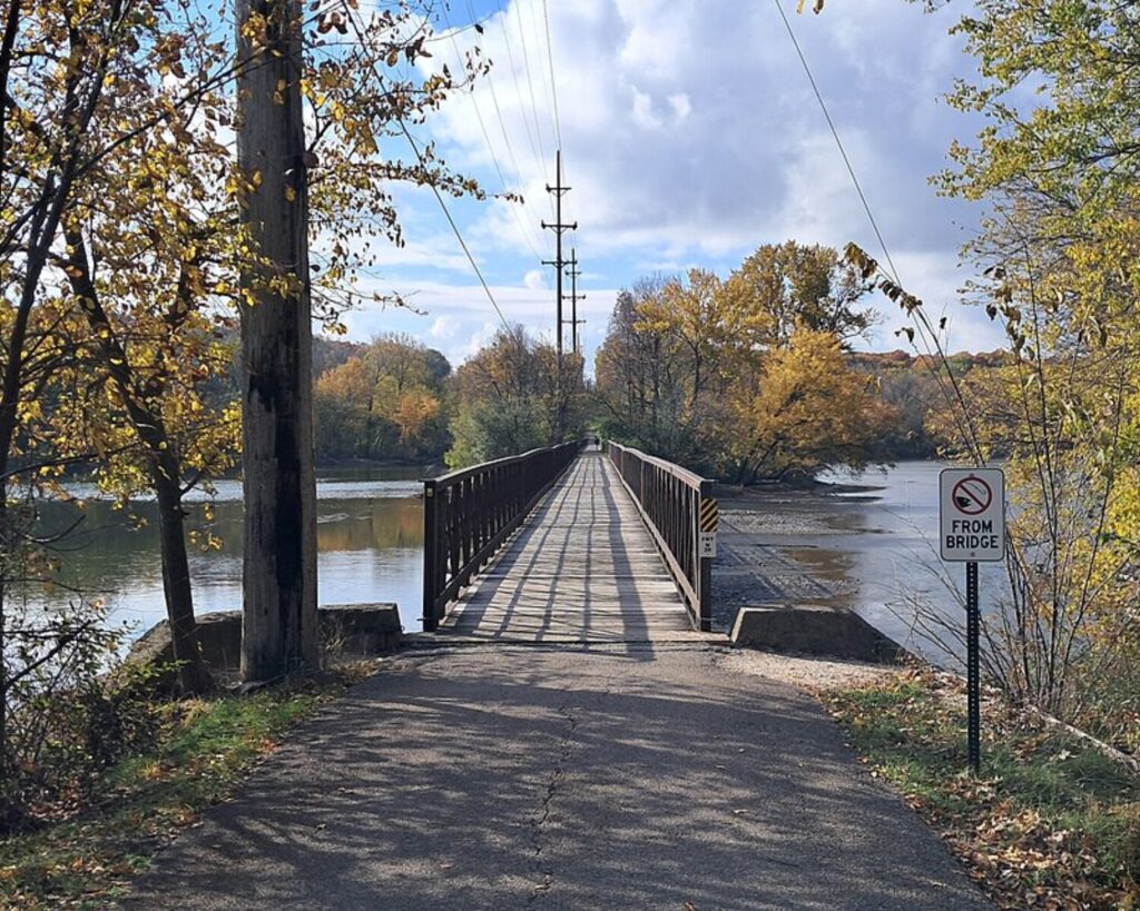 Pedestrian bridge on the Fox River Trail crossing the Fox River, surrounded by autumn trees under a partly cloudy sky.