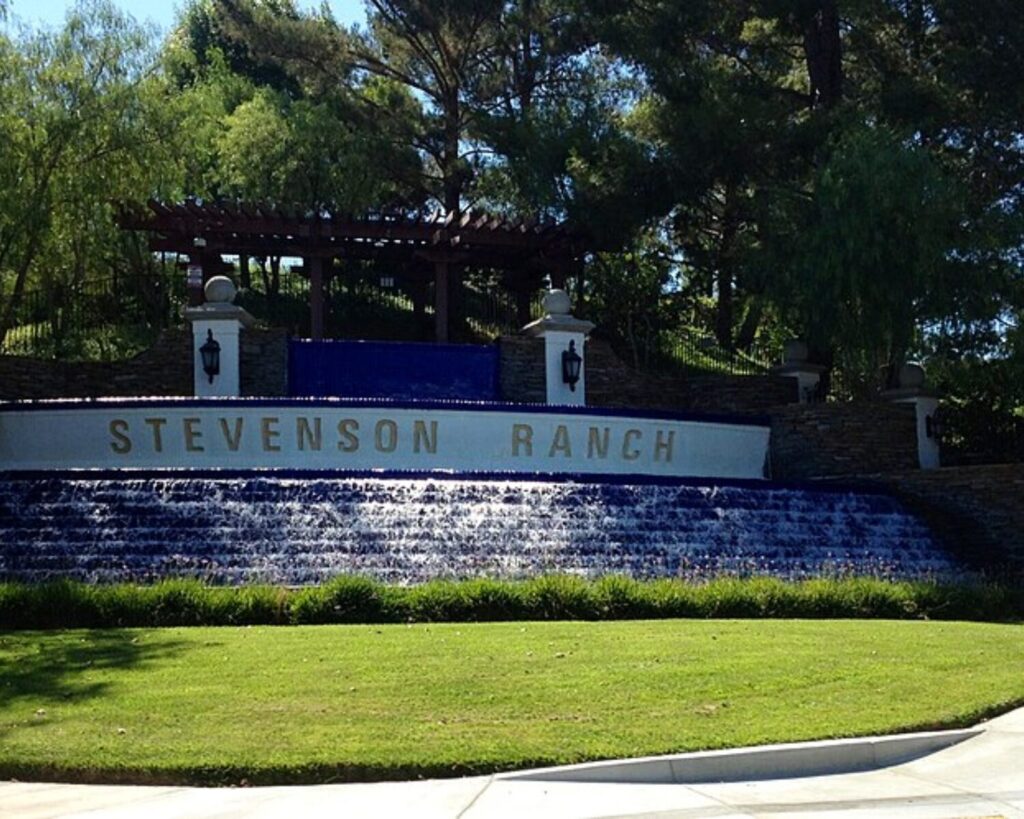 Stevenson Ranch entrance with a stepped waterfall, blue-tiled wall, and trees overhead at the entry.