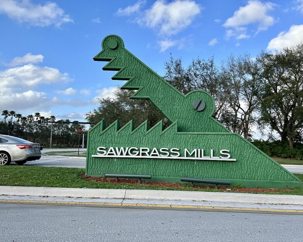 Large green Sawgrass Mills entrance sign shaped like an alligator beside a roadway in Sunrise, Florida, under a blue sky with scattered clouds.