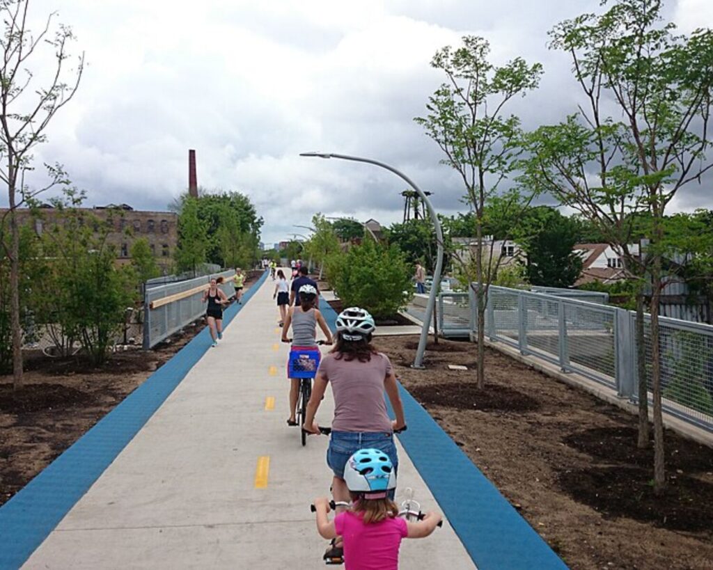 People ride bicycles on a wide elevated path with blue edges, wearing helmets, as others walk and bike in the background under a cloudy sky with trees and urban buildings nearby.