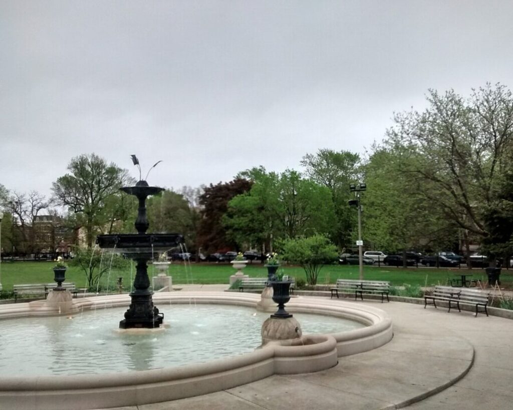Decorative fountain in Chicago’s Wicker Park featuring a circular stone basin with a historic-style tiered fountain at the center, surrounded by landscaped greenery, trees, and park benches on a sunny day.