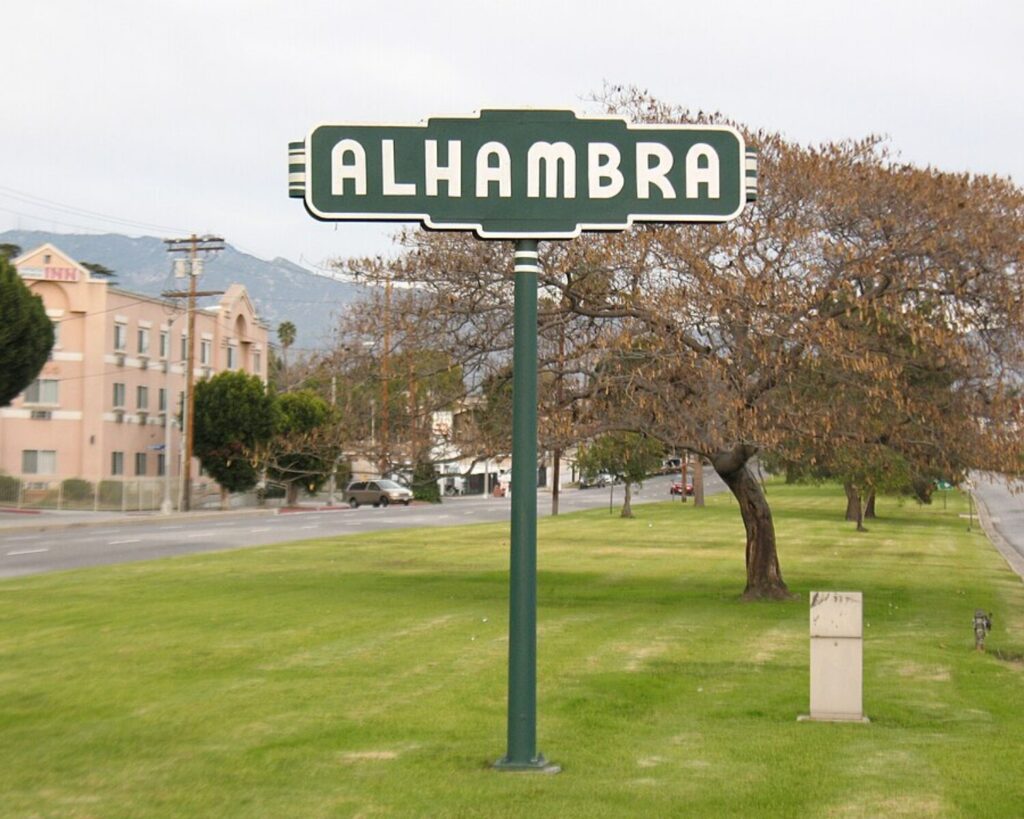 Alhambra city sign on a grassy roadside with trees, nearby buildings, and mountains in the background.