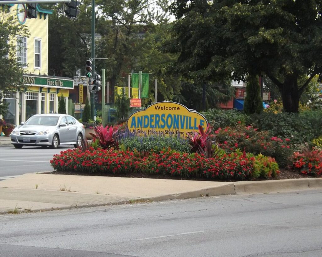 Welcome to Andersonville sign surrounded by flowers, trees, street traffic, and neighborhood storefronts.