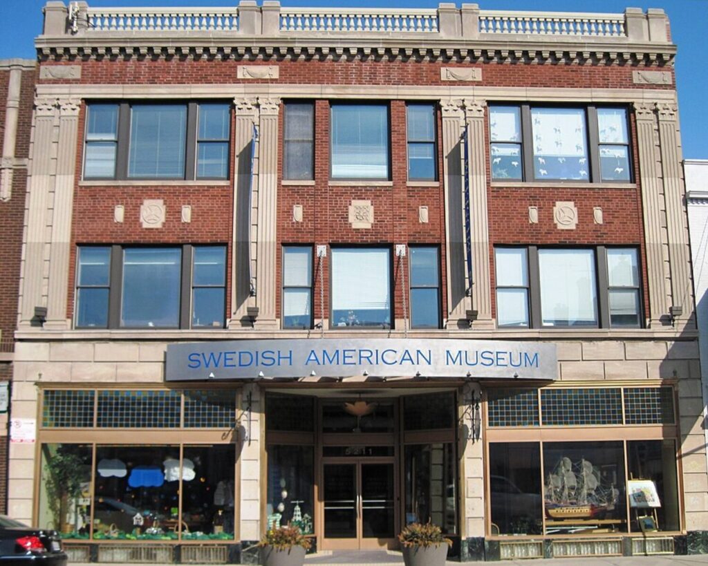 Facade of the Swedish American Museum, brick building with tall windows and decorative columns, blue sign above entrance.