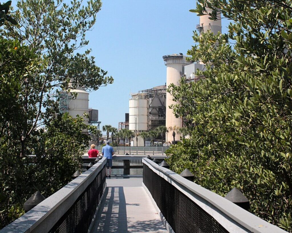 Boardwalk framed by dense trees leads to a factory with tall beige chimneys and metal structures; two people walk ahead.
