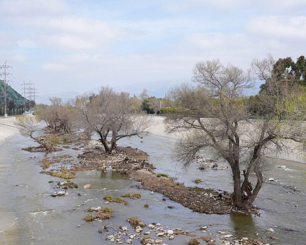 Los Angeles River in Atwater with shallow water, rocky banks, leafless trees, hills, and cloudy sky.