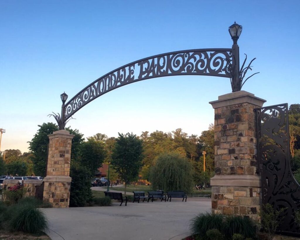 Avondale Park entrance arch with stone pillars, benches, trees, and clear blue sky.