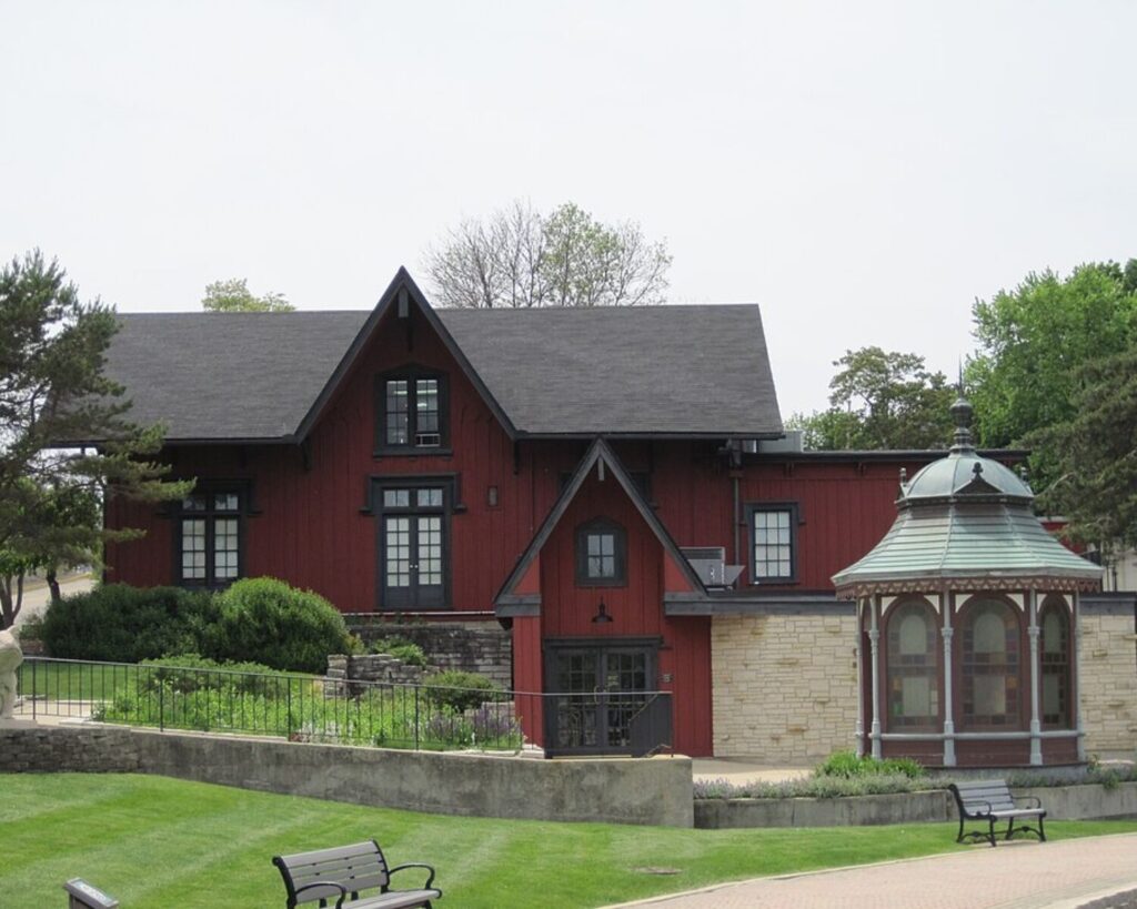 Red wooden building with black trim and steep gabled roof next to a decorative glass gazebo in a landscaped park.