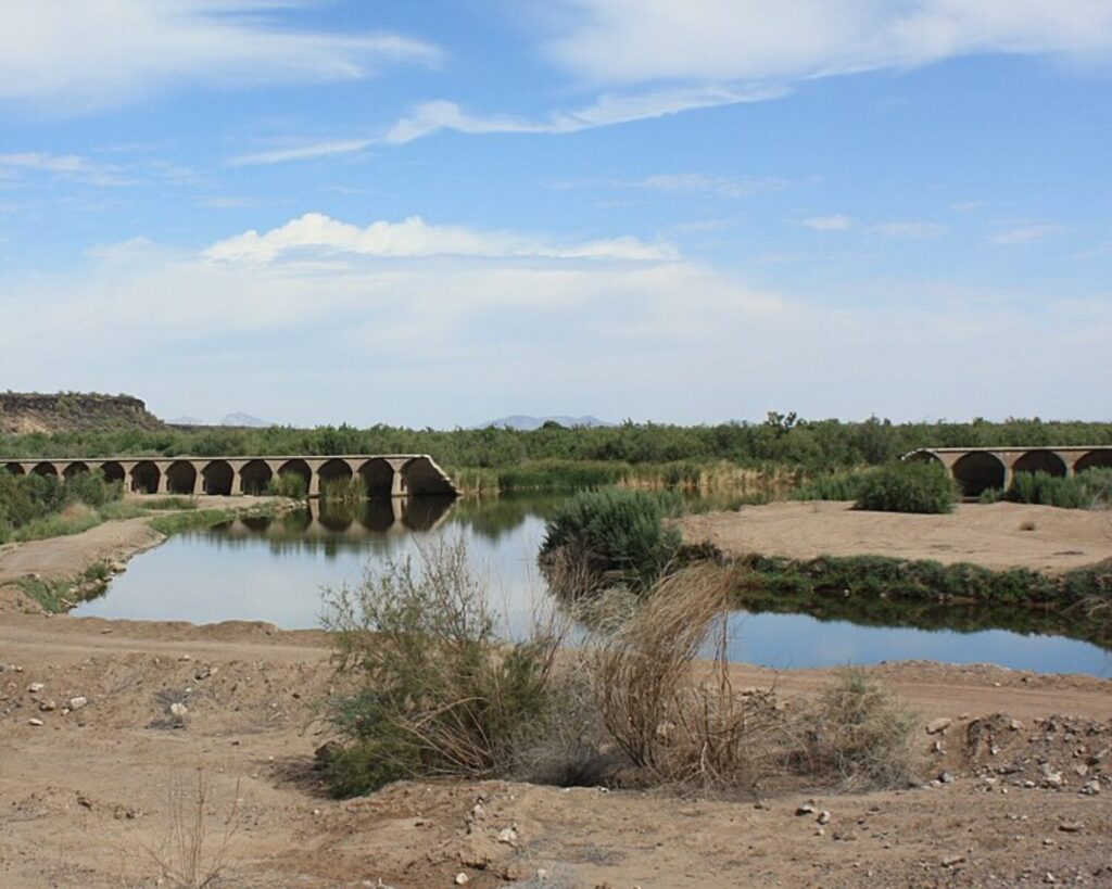 Stone arch bridge spanning a calm river in a desert landscape with blue sky and sparse vegetation.