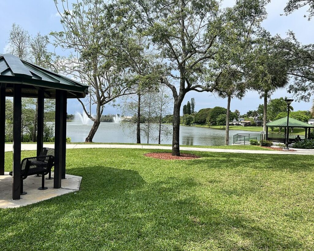 Park by a tranquil lake with a fountain, green lawn, and scattered trees, plus a shaded pavilion on the left.