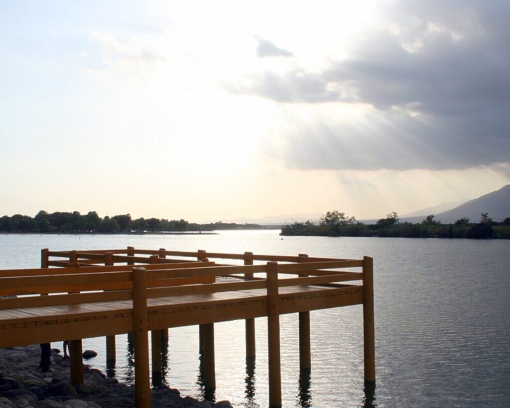 Santa Fe Dam Recreation Area with lake, wooden pier, mountains, and sun rays through clouds.