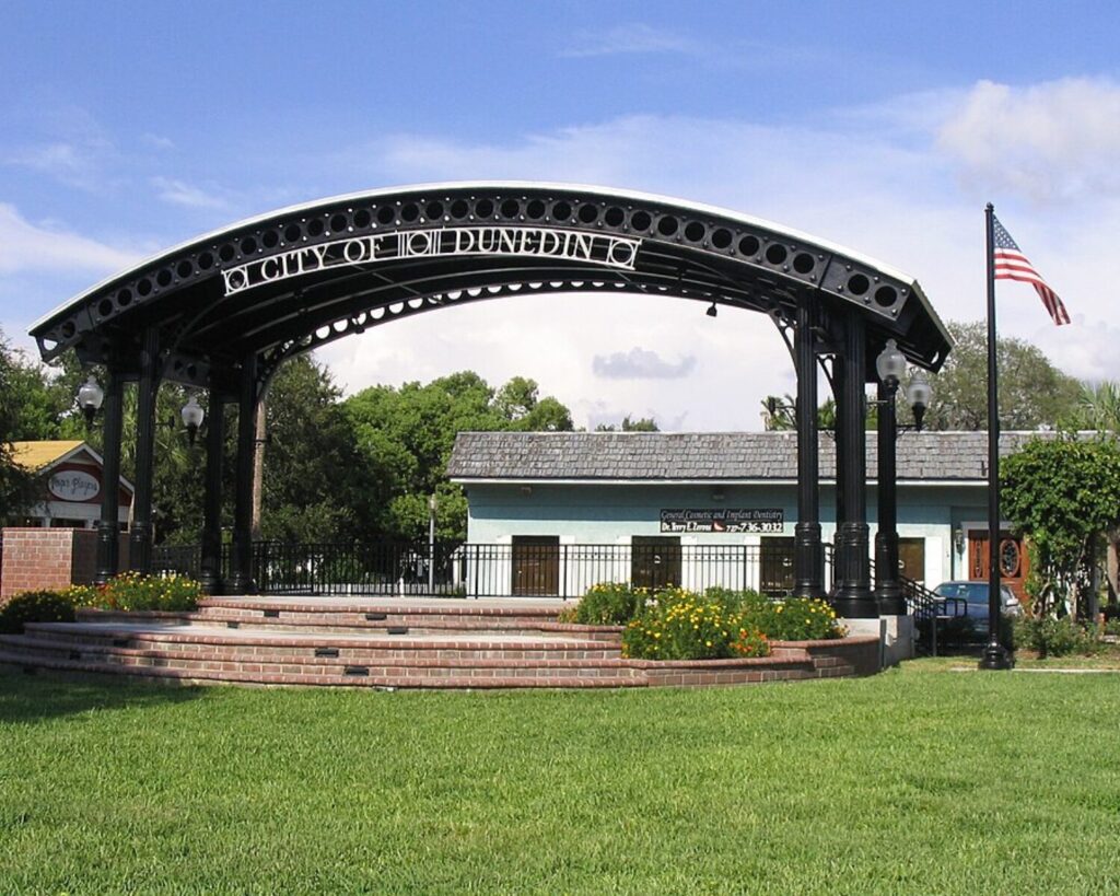 City of Dunedin bandshell stage in a grassy park with flags, trees, and a blue sky.