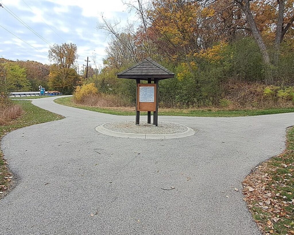 Wooden information kiosk at a circular gravel island in a park, with a map posted under a small roof, surrounded by autumn trees and a winding paved path.