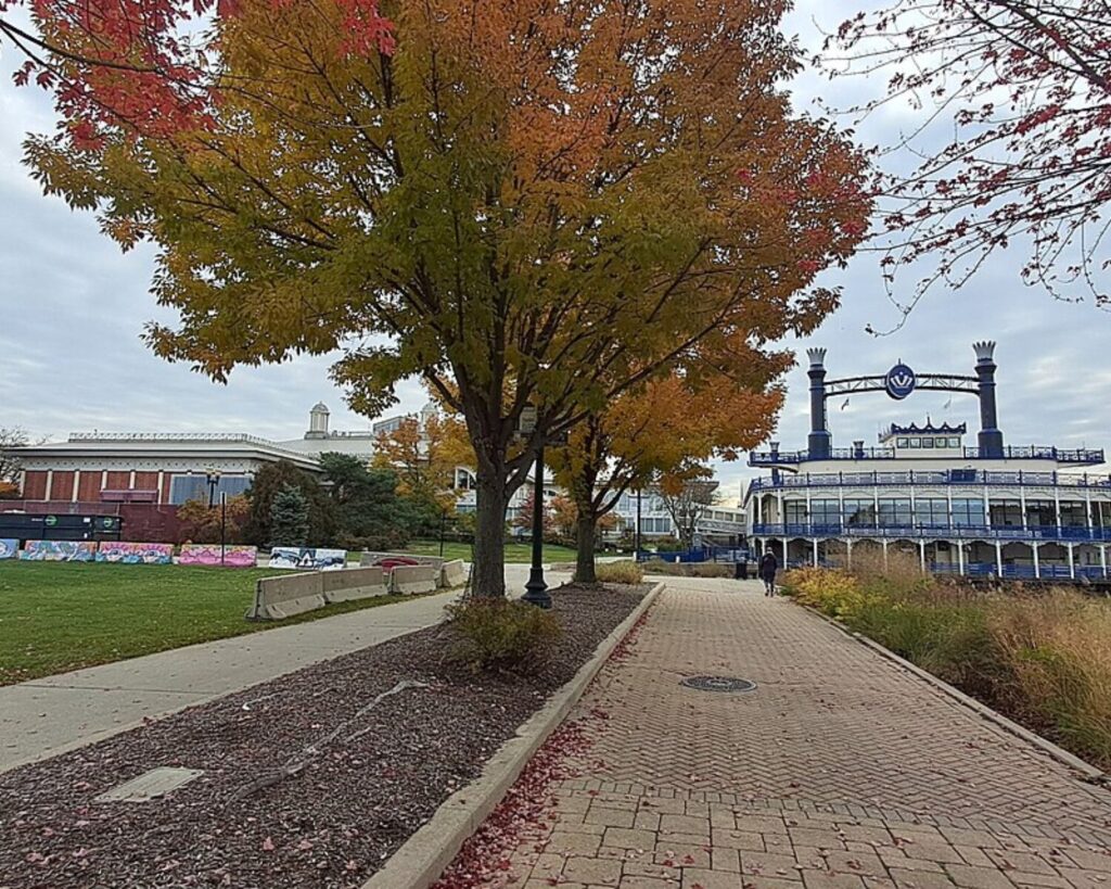 Promenade lined with autumn trees, a brick building on the left, and a blue riverside structure to the right, with a person walking in the distance.