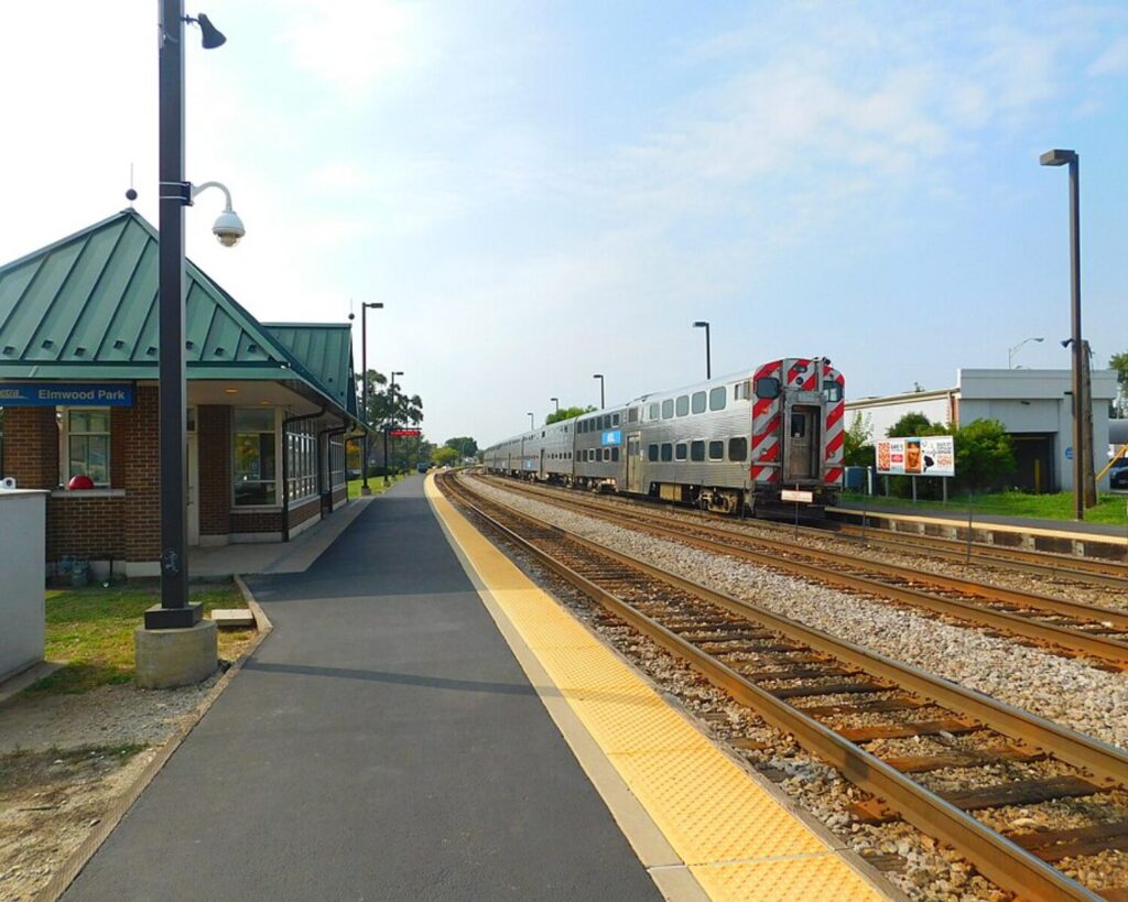Elmwood Park Metra station platform with train tracks, a parked train, and a small station building under a bright sky.