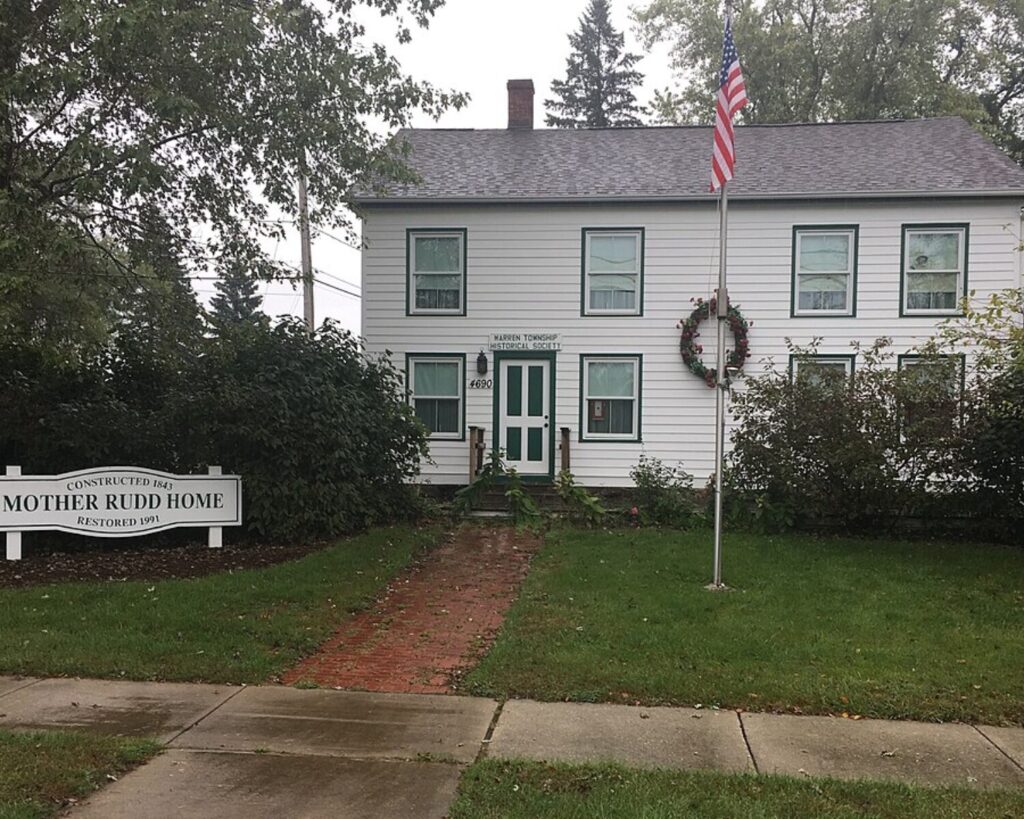 Mother Rudd Home exterior with white historic house, sign, flagpole, lawn, and trees.