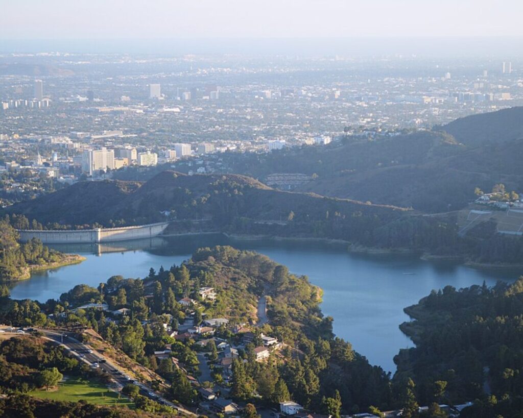 Aerial view of a lake with a dam, surrounded by forested hills and a distant city skyline in the background.