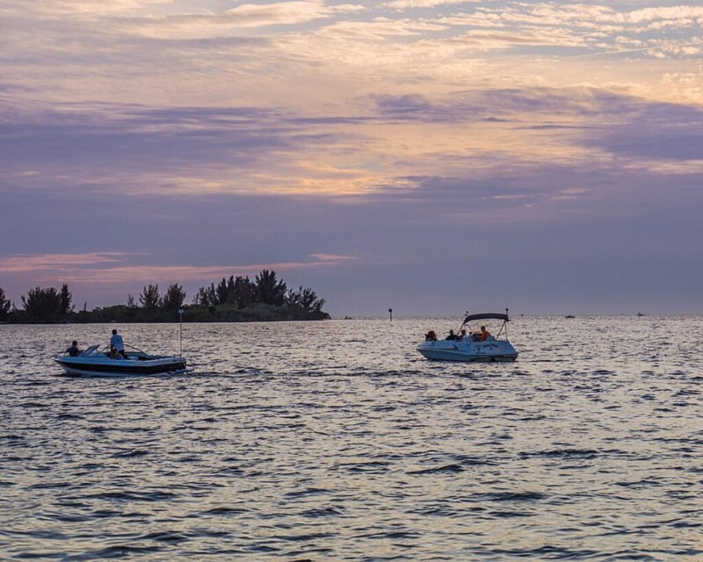 Two small boats glide on calm water at sunset with a tree-covered island in the distance.