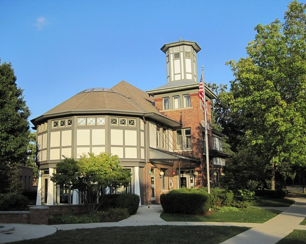 Morton Grove Historical Museum with a green historic house, parking lot, hedges, tall trees, and cloudy sky.