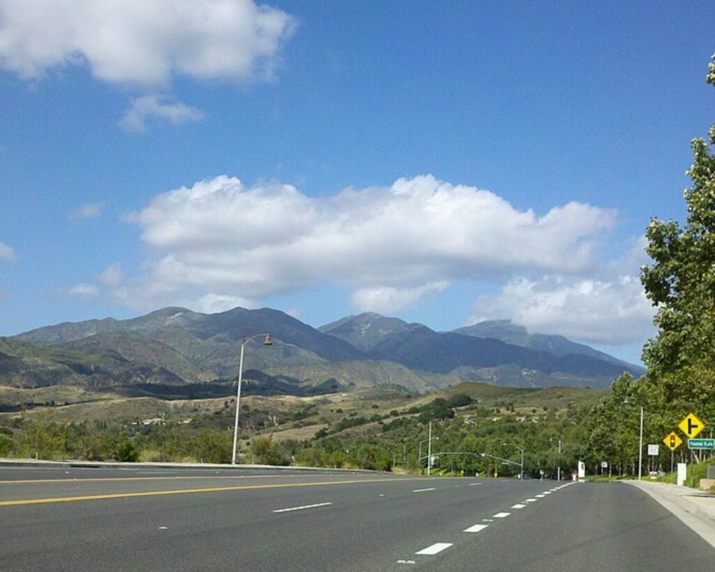Open highway with multiple lanes and mountains in the distance under a blue sky with fluffy clouds; road signs visible on the right.
