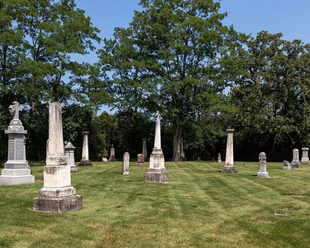 A sunny cemetery with tall stone obelisks and memorials scattered over neatly mowed grass, framed by green trees and a blue sky in the background.