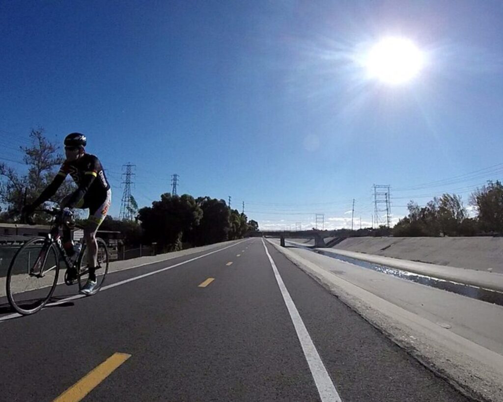Cyclist in helmet and dark cycling kit riding in a dedicated bike lane on a sunny day.