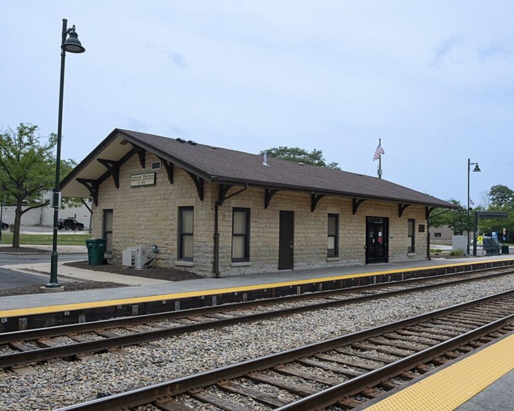 Lemont Station building beside railroad tracks with platform, lamps, and clear blue sky.