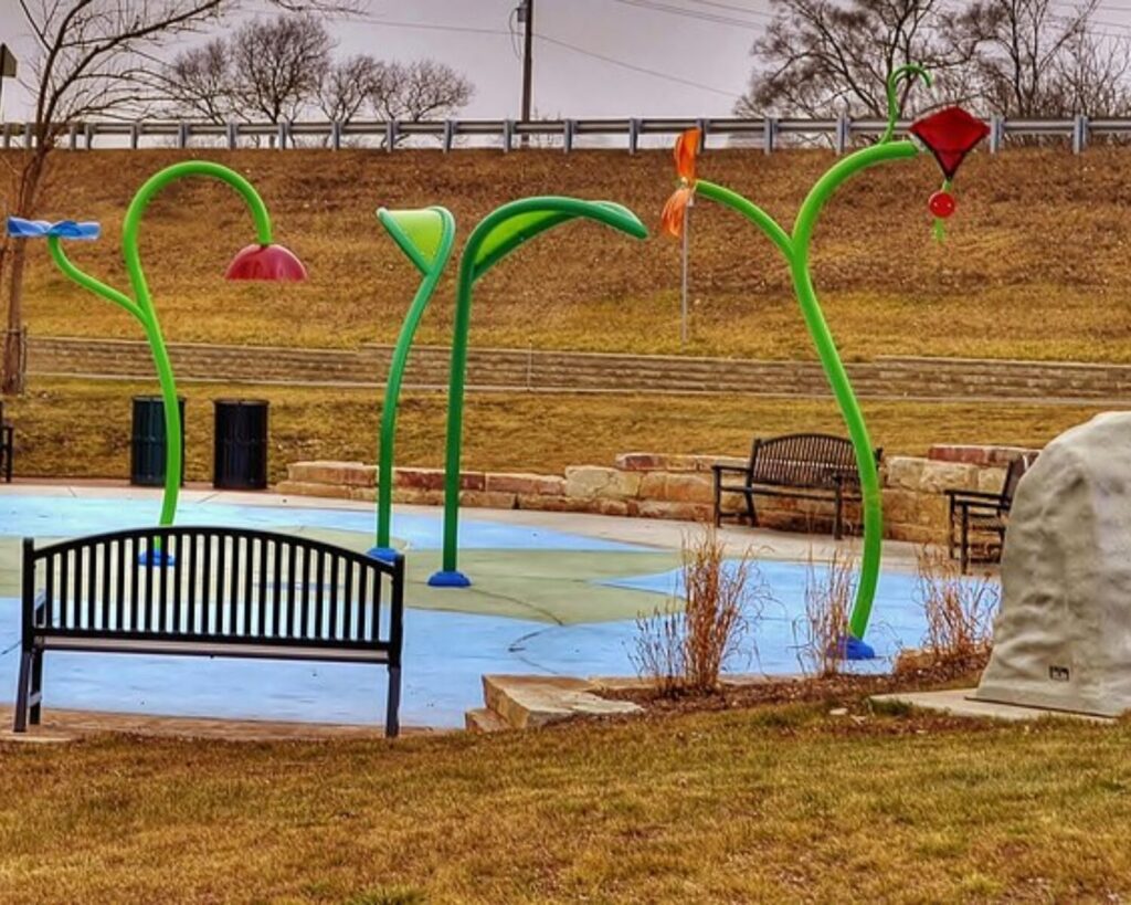 Bright green vine-like water play structures with flower-shaped spray heads at a splash pad, with benches in the background.