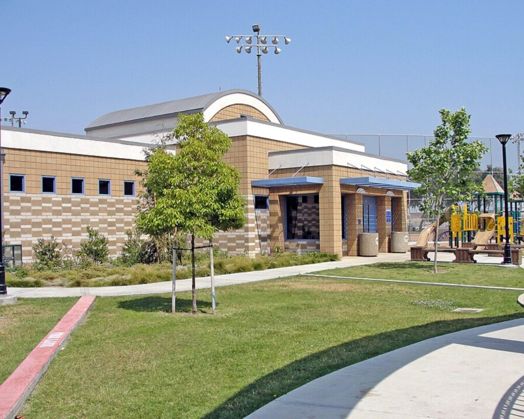 Maywood Park Aquatic Center building with green lawn, playground equipment, and a clear blue sky.